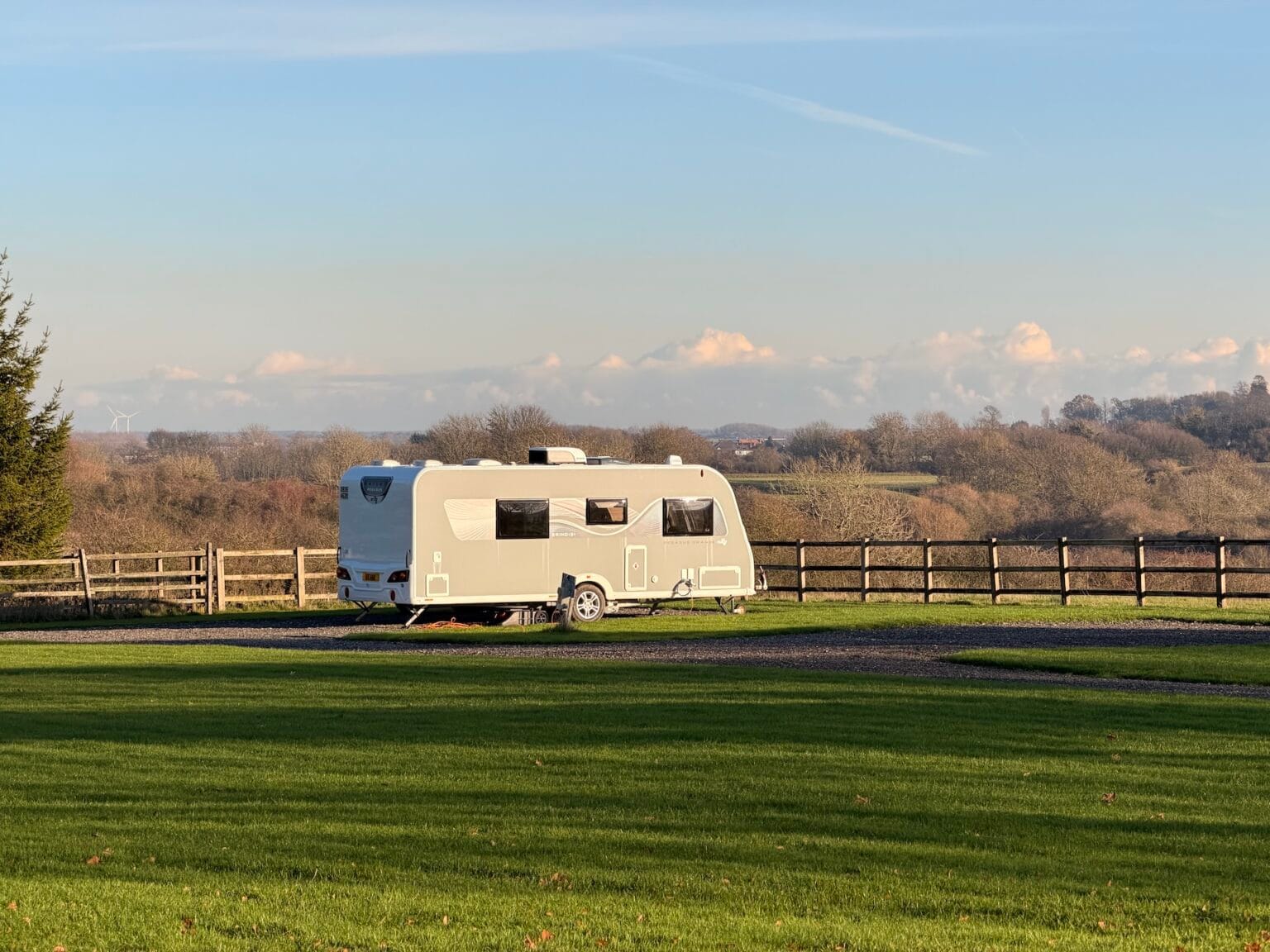 A modern grey-coloured caravan parked on a spacious gravel pitch overlooking rolling countryside under a clear blue sky.